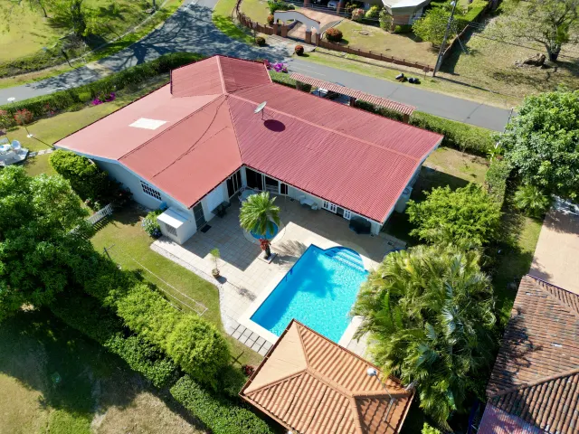 An aerial view shows a single-story house with a reddish metal roof, a backyard pool, and a tiled patio surrounded by green lawns.