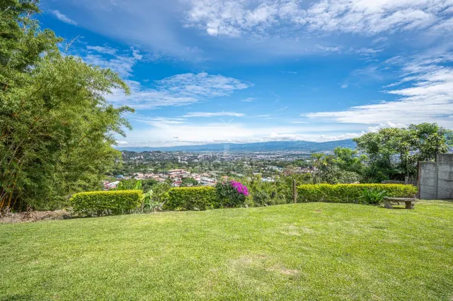 An open green lawn with trimmed hedges frames a wide-angle view of a distant urban valley and mountains under a blue sky with scattered clouds.