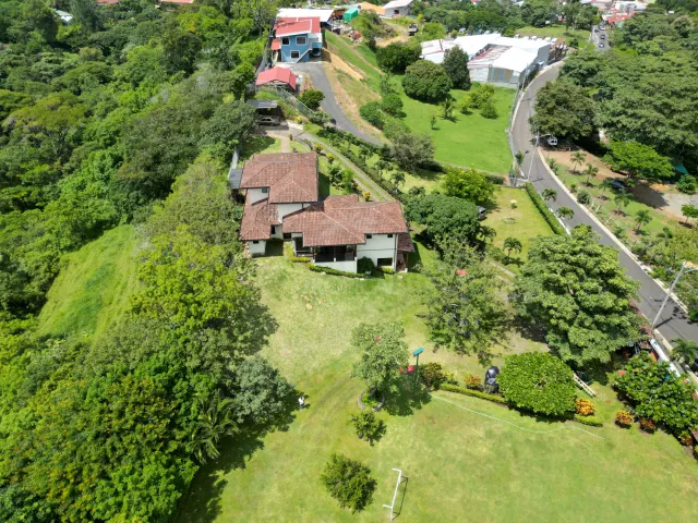 High angle shot showing a house with a tiled roof surrounded by green grass and trees, next to a road.