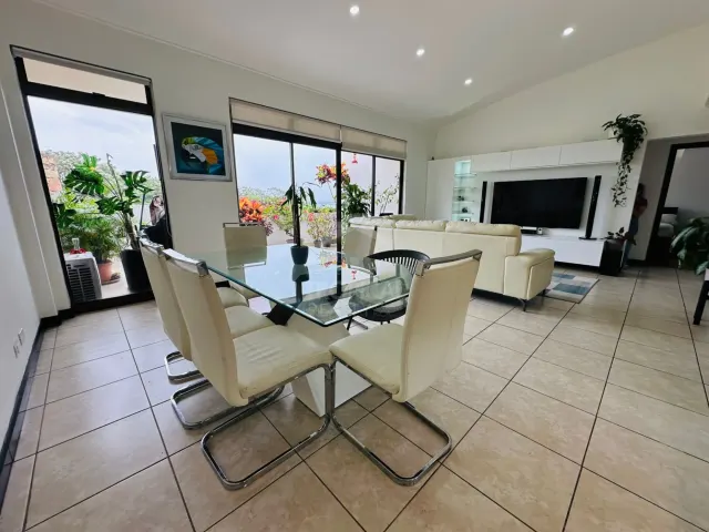 Living and dining area featuring a glass-topped table with white chairs, tile floors, a white leather sofa, and a television unit against the wall. Large windows open to a plant-filled balcony.