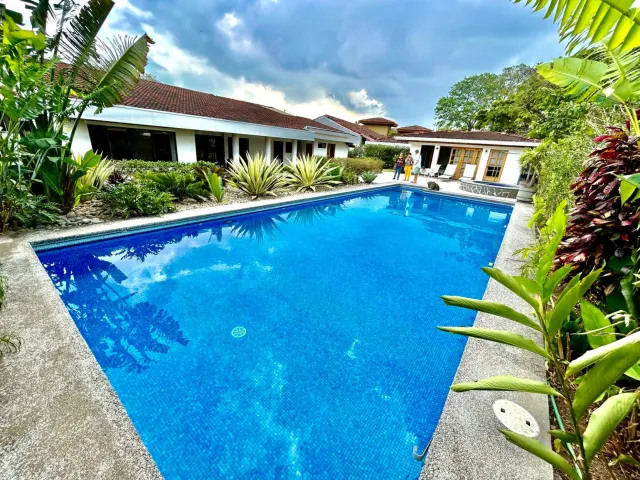 A rectangular swimming pool with blue tile interior is surrounded by tropical plants and single-story white buildings with tile roofs under a cloudy sky.