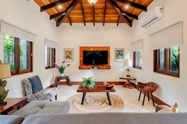 A living room featuring a vaulted wood-beam ceiling, white walls, and tile flooring. Furnishings include a sofa, armchairs, a coffee table on a round rug, and a wall-mounted television.
