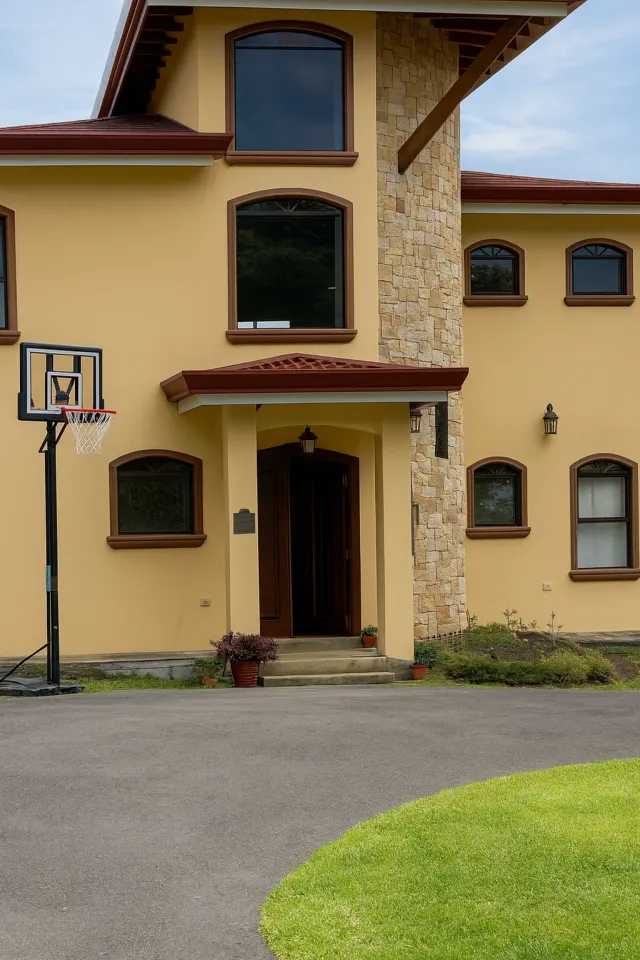 Exterior view of a yellow two-story building featuring dark brown window frames, a stone pillar, and a portable basketball hoop on the paved driveway.