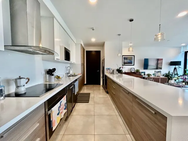 Kitchen featuring a long white quartz countertop island, wooden base cabinets, stainless steel appliances, and tile flooring. A living area is visible in the background.