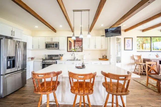 Kitchen featuring a white central island with three wooden bar stools, white cabinetry, stainless steel appliances, and exposed wooden ceiling beams.