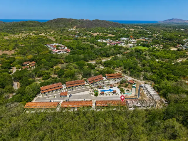 Aerial view showing multiple buildings with red tile roofs, parking areas, and a swimming pool surrounded by green trees and distant hills.