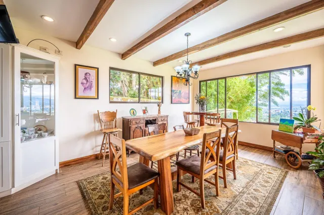 Dining room featuring a wooden table with chairs, wood-beamed ceiling, glass-front display cabinet, and large windows with garden views.