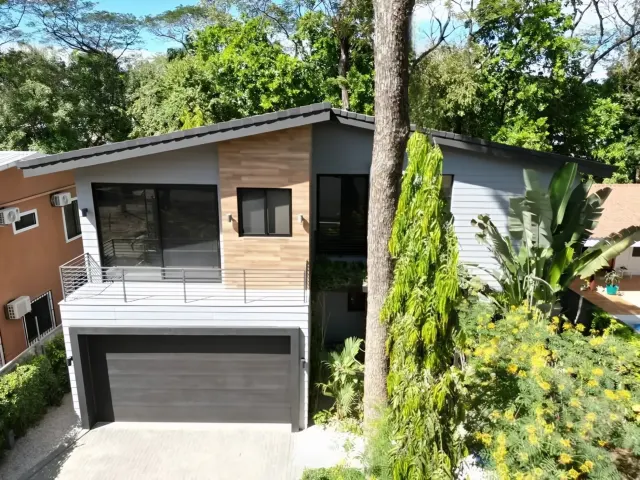 Two-story modern house exterior with gray siding, a dark garage door, and a second-floor balcony, surrounded by trees and foliage.