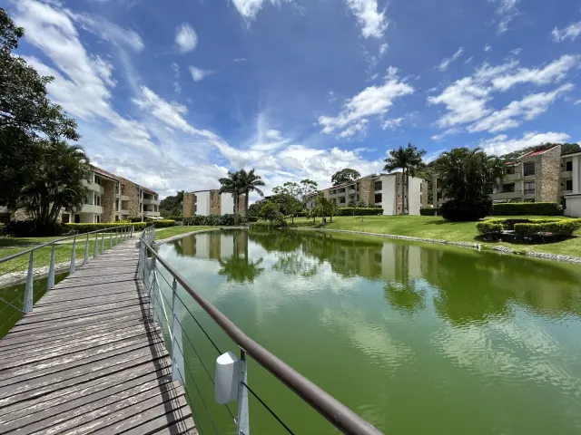 A wooden pedestrian bridge with metal railings curves over a green pond, surrounded by apartment buildings and grassy lawns under a blue sky.