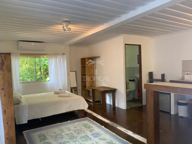 Studio room with a bed, a window with greenery outside, wooden furniture, and a doorway leading to a bathroom.