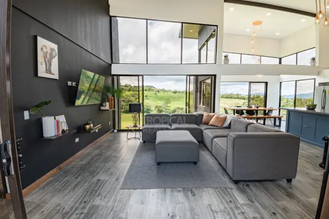 Open-plan living room featuring a grey sectional sofa, grey wood-look tile floors, and a double-height ceiling with floor-to-ceiling windows showing a green landscape.