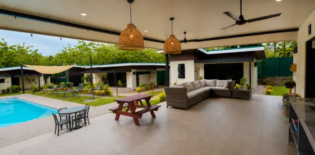 A tiled covered patio features a large wicker sectional sofa, a wooden picnic table, and hanging woven lamps, overlooking a swimming pool and courtyard.