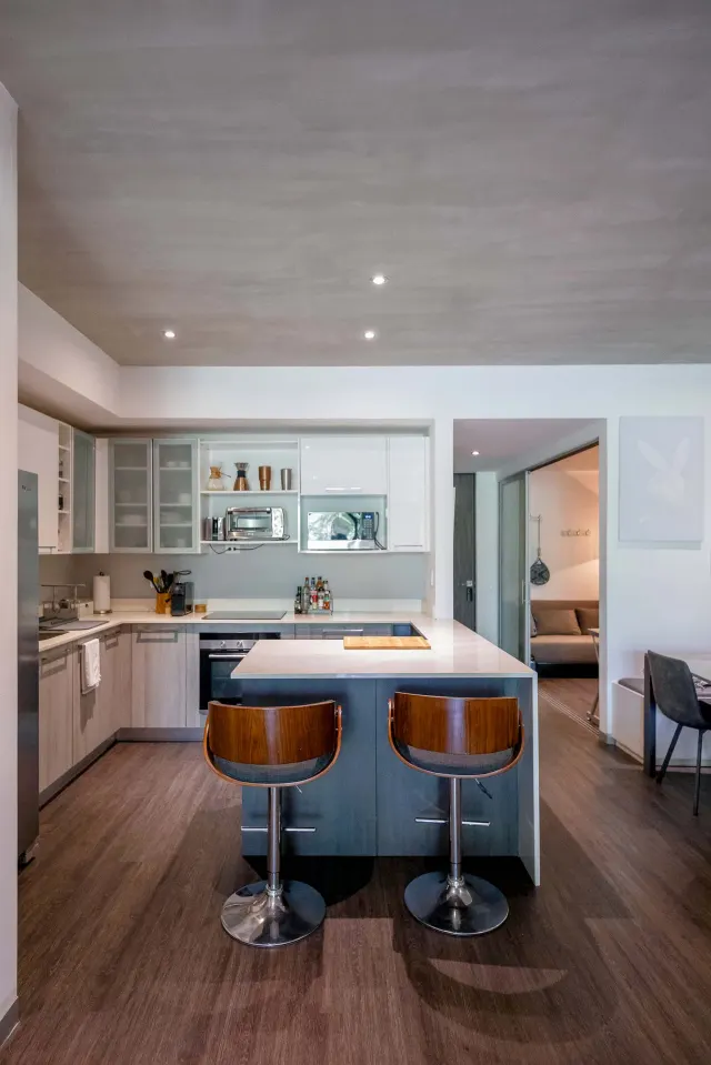 Kitchen with wooden laminate floors, a central island, two bar stools, white and glass cabinets, and built-in appliances.