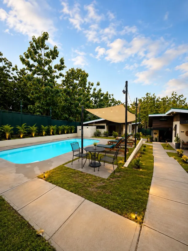 Outdoor area with a rectangular swimming pool, a table with four chairs under a shade sail, lounge chairs, and concrete walkways flanked by grass and lights.