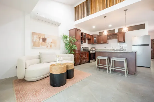 Open-plan living area with a cream sofa, pink rug, and two small round tables. An adjoining kitchen features dark wood cabinets and a breakfast bar with two stools.