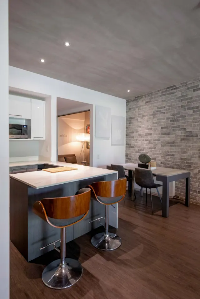 Kitchen counter with two wooden bar stools facing an adjacent dining table and a grey brick wall.