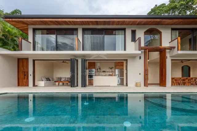 Two-story home facade with a swimming pool in the foreground, open glass folding doors, and wooden ceiling details.