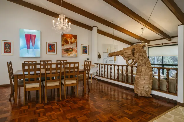Dining area featuring a wooden table with chairs, parquet floors, exposed ceiling beams, and artwork on the wall. A large woven sculpture stands near a wooden railing.