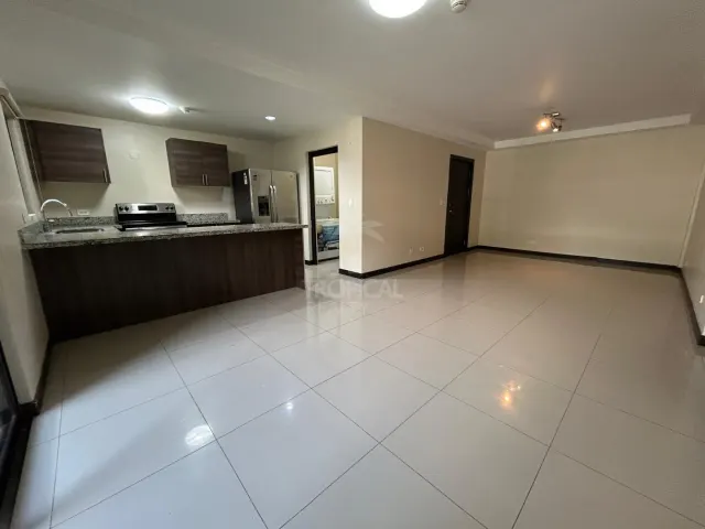 An open indoor space featuring a kitchen with dark cabinetry, granite countertops, and a stainless steel refrigerator. The adjoining room has white tile floors.