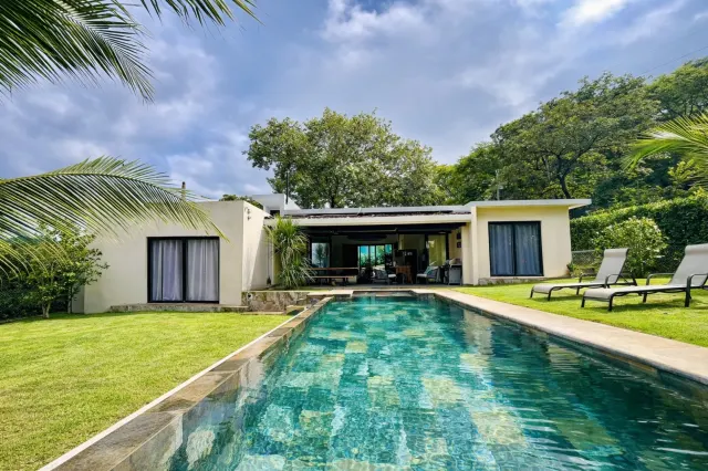 Exterior of a single-story house featuring a long lap pool in the foreground, surrounded by a green lawn and mature trees.