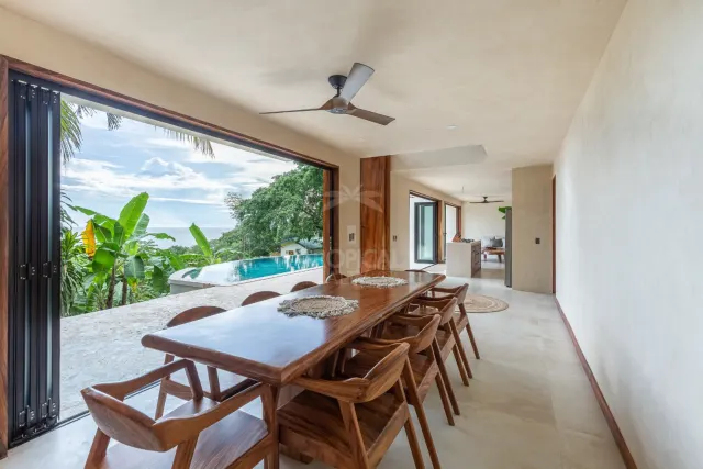 An open covered terrace featuring a long wooden dining table with matching chairs, ceiling fan, and a view of an outdoor pool and the sea through a large opening.