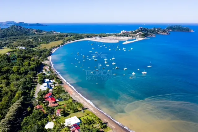 High angle view of a coastal bay with numerous anchored boats, a sandy beach, and surrounding vegetation under a clear sky.