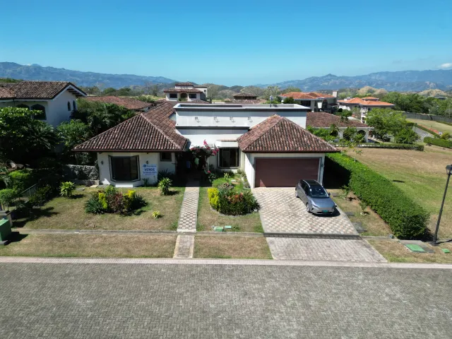 Aerial view of a white one-story house with a brown tiled roof, a paved driveway, and mountains in the distance under a blue sky.