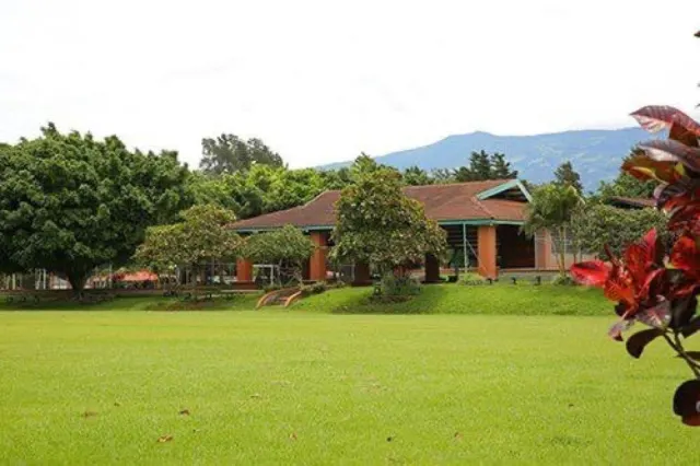 An expansive green lawn with a low single-story brick building and tiled roof in the background, surrounded by mature trees and distant mountains.