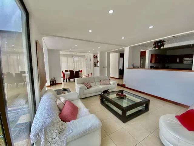 Living area with white leather sofas, a glass-topped wooden coffee table, tile flooring, and recessed ceiling lights. A dining area and kitchen are visible in the background.