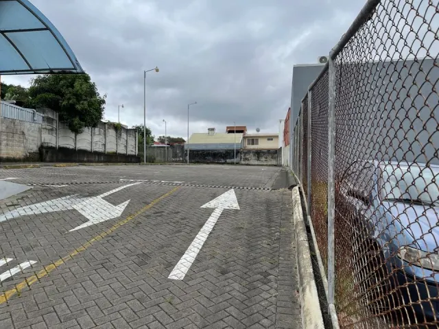An empty paved parking lot with painted directional arrows on the ground, enclosed by a chain-link fence and concrete walls under an overcast sky.