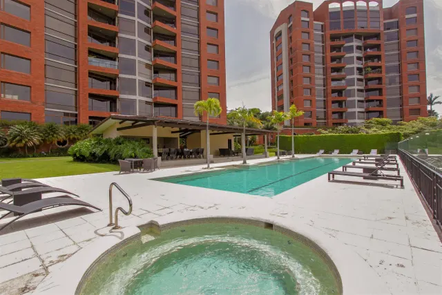 An outdoor swimming pool and circular hot tub on a tiled deck, surrounded by lounge chairs, palm trees, and multi-story red brick buildings.
