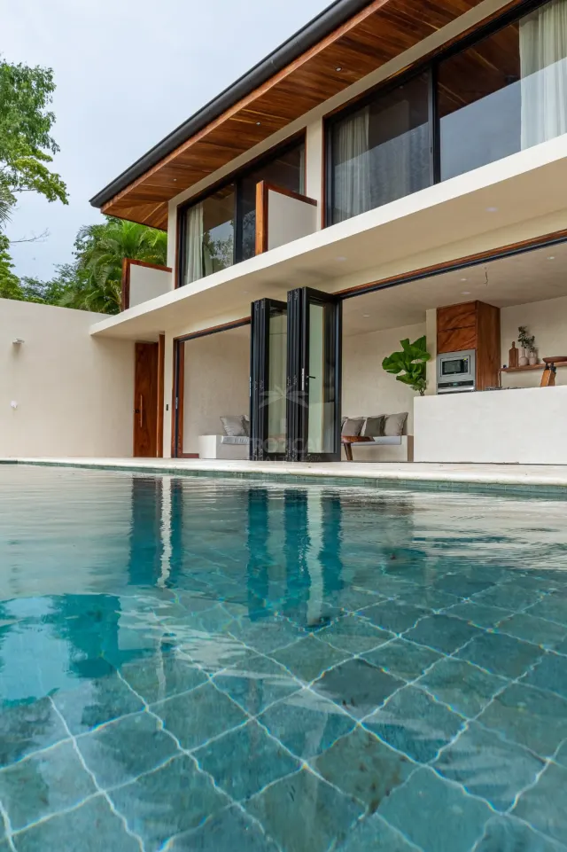 Two-story house exterior with a swimming pool in the foreground, wooden eaves, and folding glass doors opening to an indoor living area.