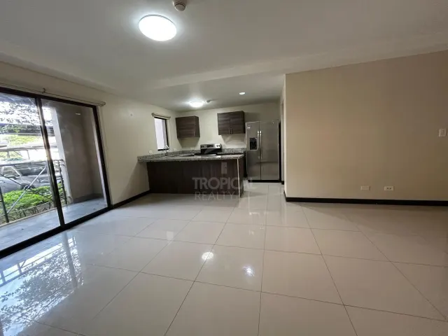 Open living area with glossy tile floors leading to a kitchen featuring dark wooden cabinets, a breakfast bar, and stainless steel appliances. Sliding glass doors open to a covered patio.