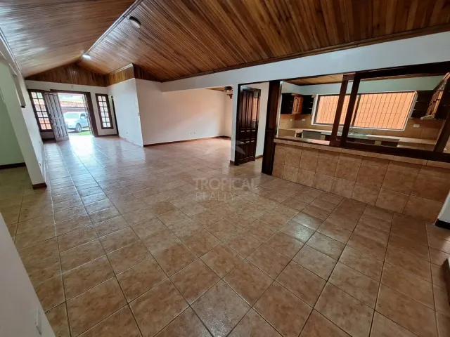Empty living room with light brown tile flooring, vaulted wood-plank ceiling, and a kitchen service window on the right.