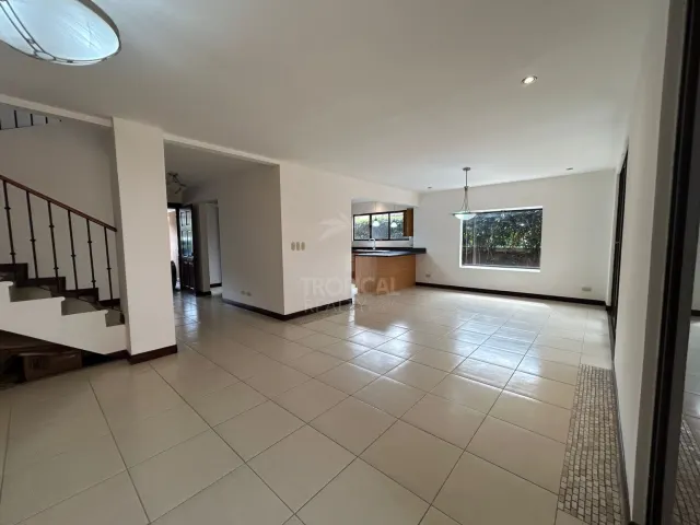Empty living space featuring light-colored tile floors, a staircase with a dark railing, and a view into the kitchen.
