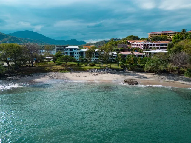 An aerial view showing buildings along a sandy beach with green vegetation, mountains in the background, and the ocean in the foreground.